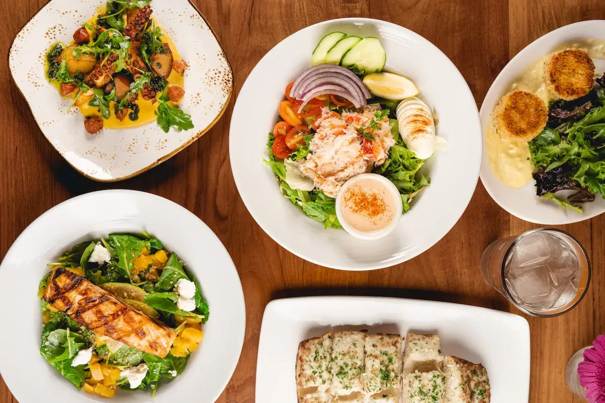 Five plates on a wooden table including a Louie Salad, Octopus Appetizer, Cheesy Garlic Bread, Kale Salad with Salmon and Crab Cakes.