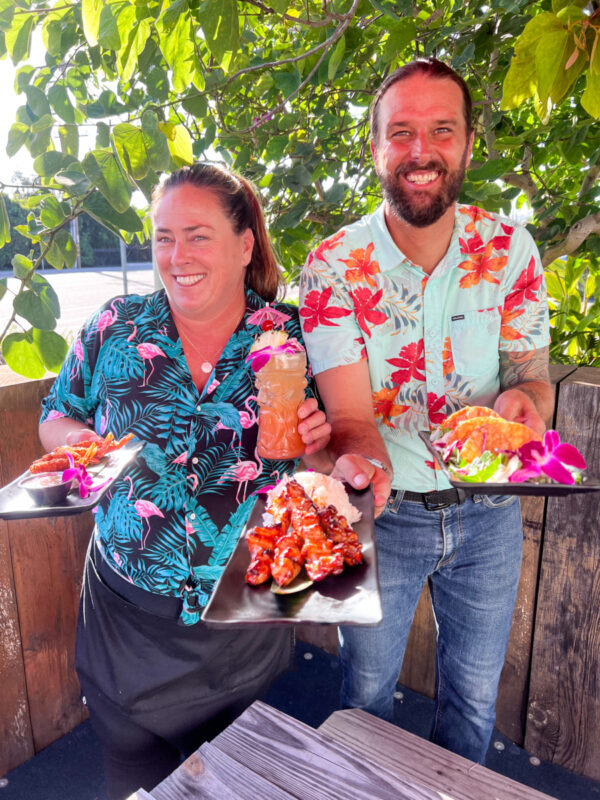 A male and female Fish Market employee dressed in Hawaiian shirts with a Mai Tai and a few appetizers in their hands. They are on the patio with trees in the background.